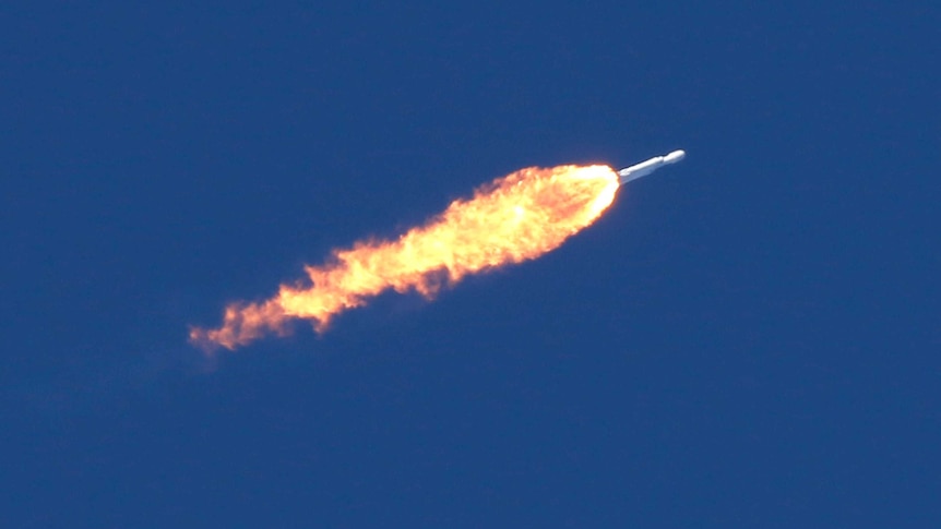 Flames spew out from behind a rocket set against a deep blue sky.
