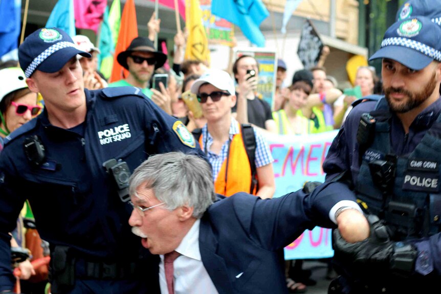 Two police men drag an elderly man in a suit as protesters look on in the background.
