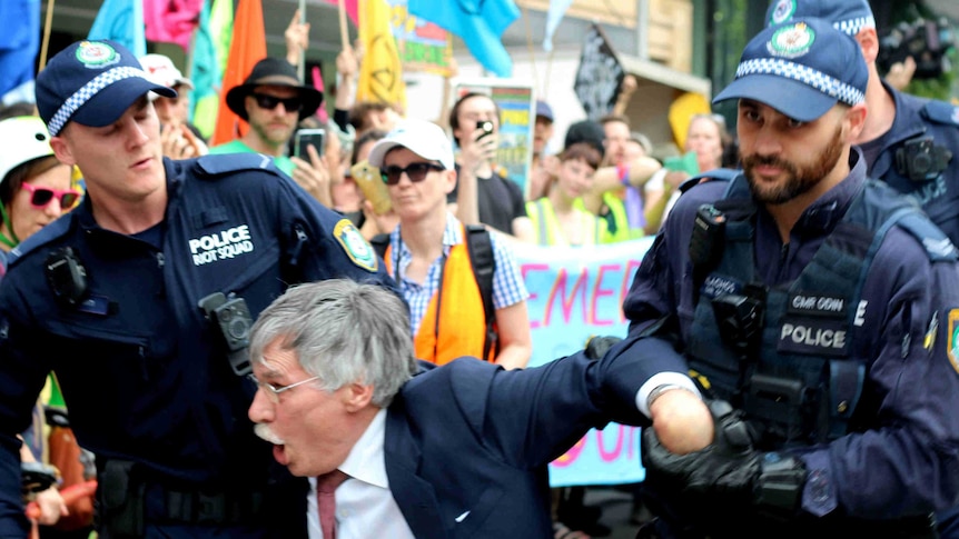 Two police men drag an elderly man in a suit as protesters look on in the background.