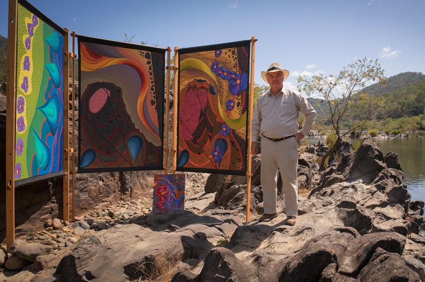 Dr John Jackson stands on rocks beside the Clarence River with a gallery of four of his paintings.