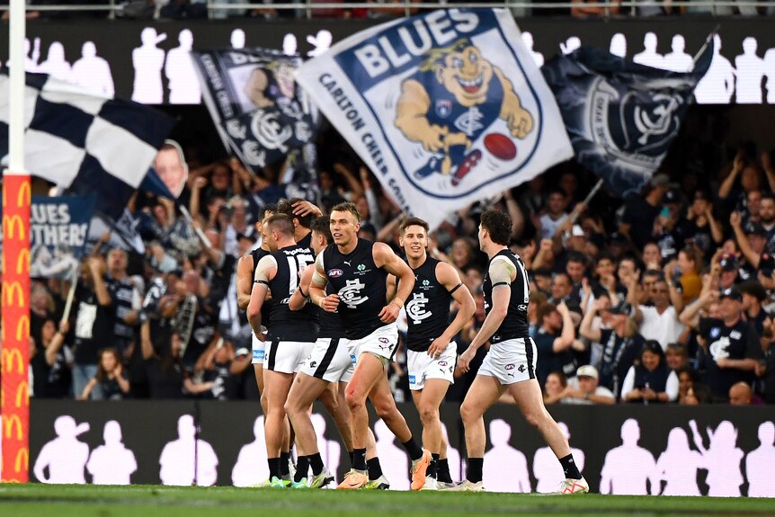 A group of Carlton AFL players run back to the centre after a goal, while fans in the stands hold a big flag in the background.