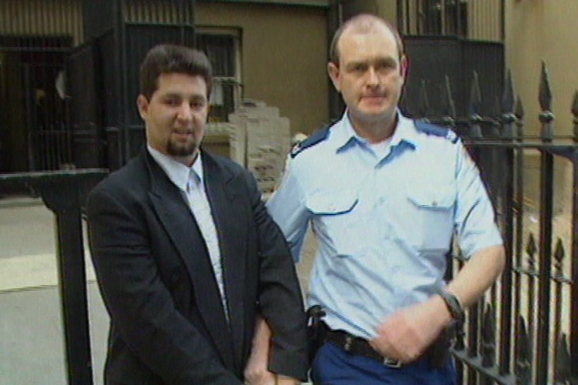 A young, dark-haired man in a suit is led by a police officer from a court building.