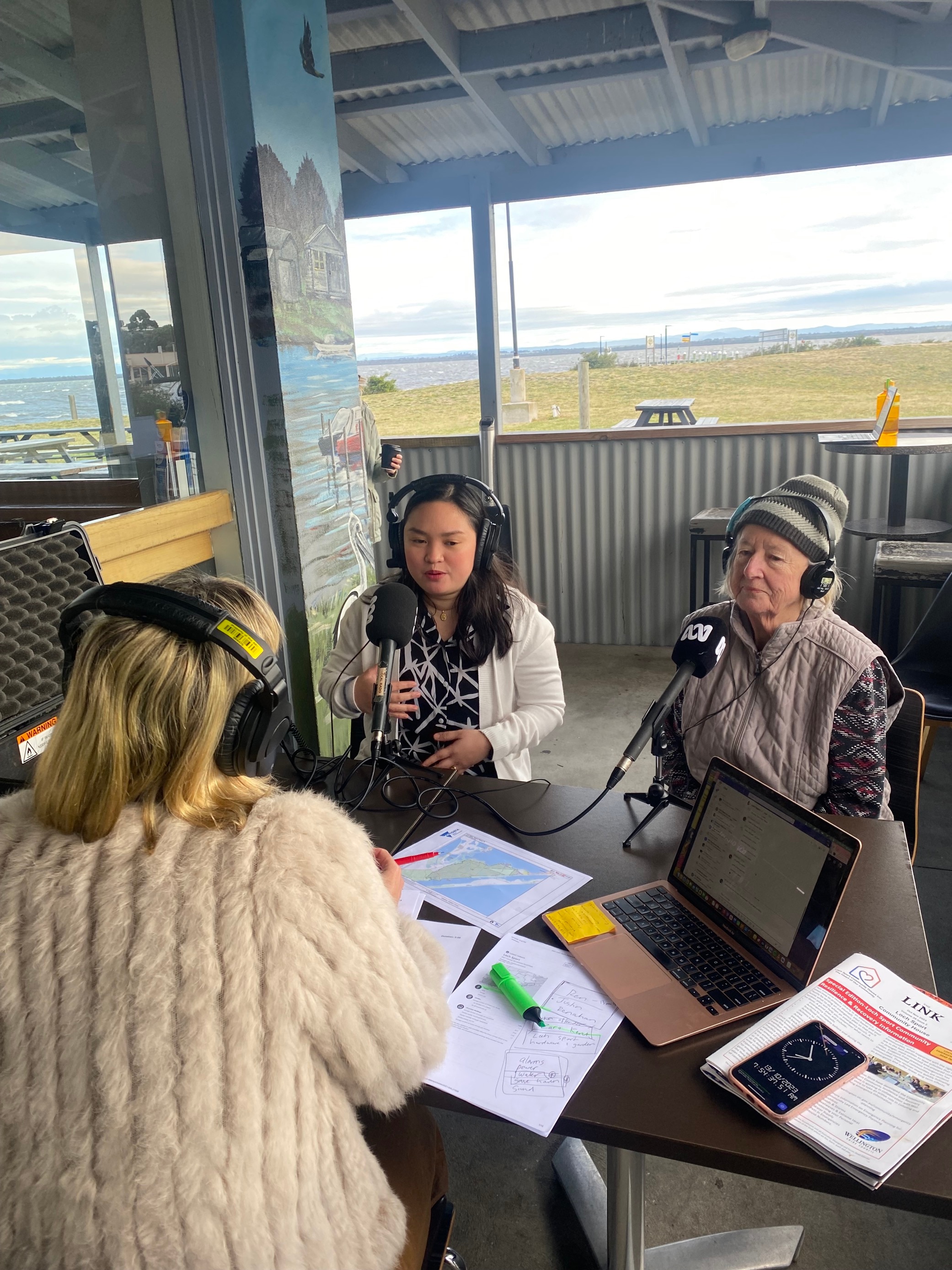 Three women with headphones on sitting at an outside table in front of ABC microphones.