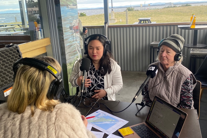 Three women with headphones on sitting at an outside table in front of ABC microphones.