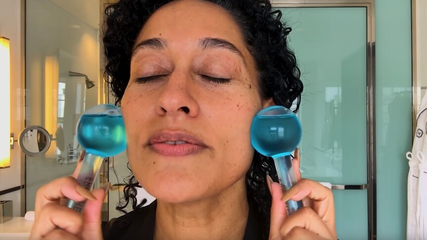 A woman presses two round glass face-massagers to her cheeks to depict chasing unrealistic beauty standards.