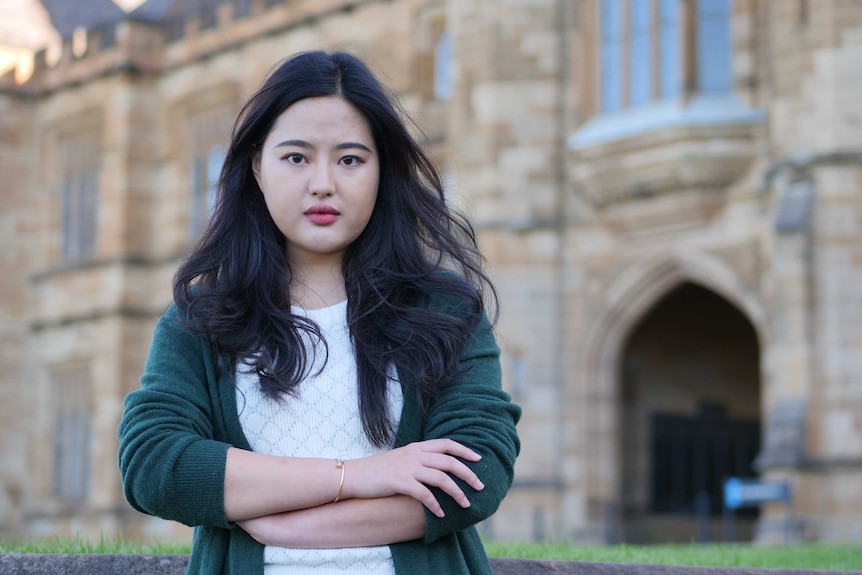 A young woman of Asian background stands with her arms crossed in front of a historic-looking building.