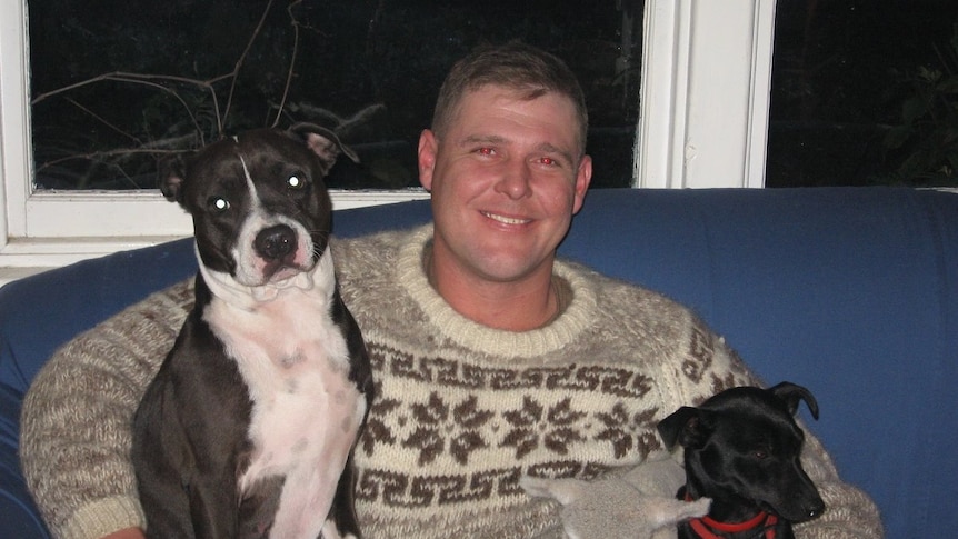 A smiling man sits on a couch with his two dogs and a lamb.