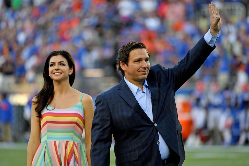 A man in a suit waves while walking with a brunette in a brightly coloured dress