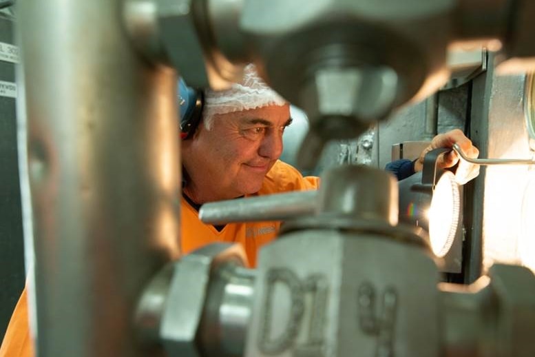 A man in a hair net and orange overalls is seen through gaps in machinery