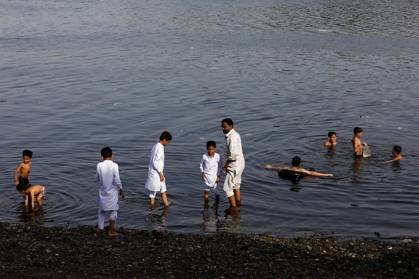 People, some dressed in white cloth others shirtless, swim or wade in shallow dark waters with dark sands and stones