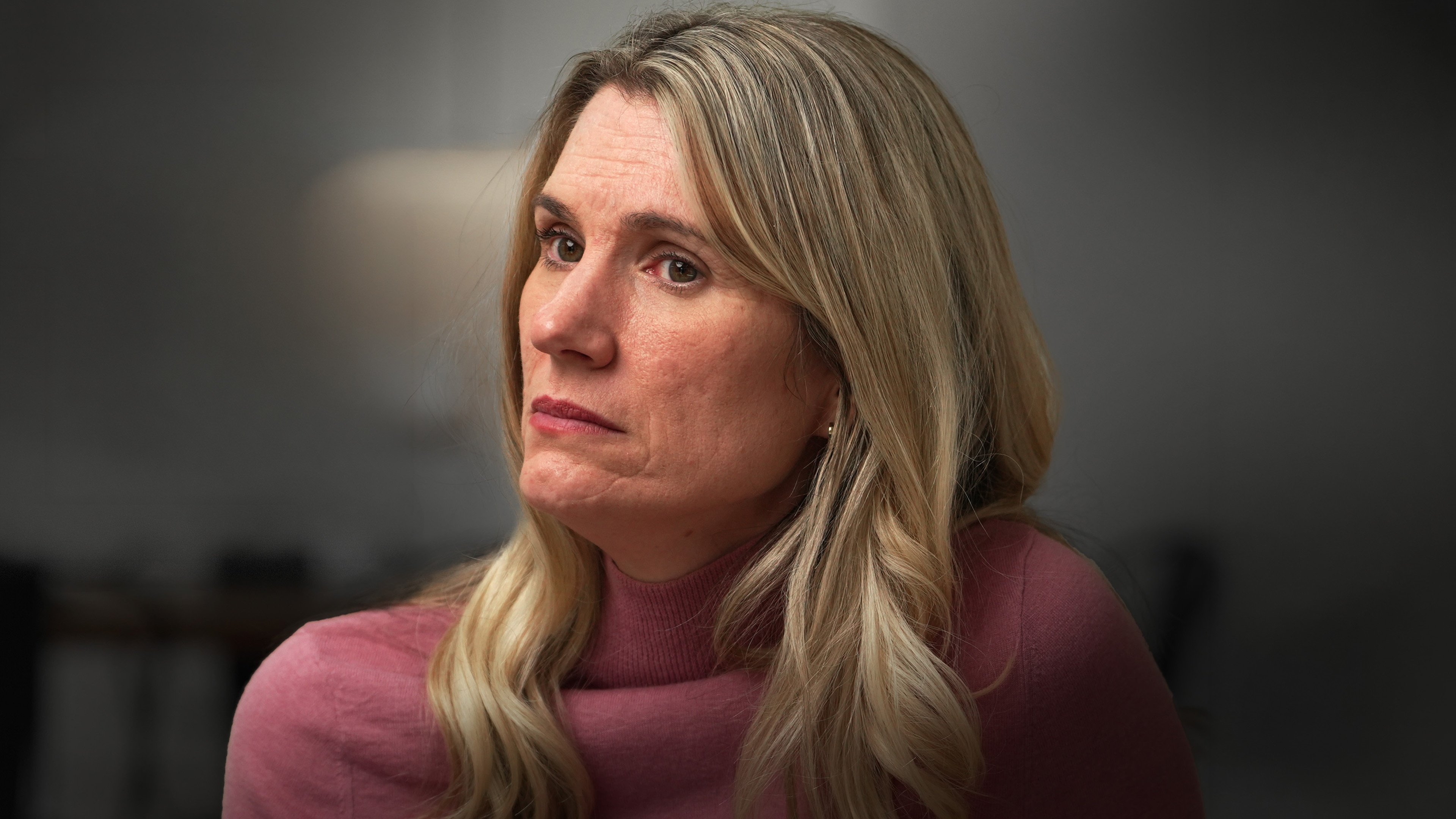 Woman with shoulder-length blonde hair wears a pink sweater with a serious expression on her face.