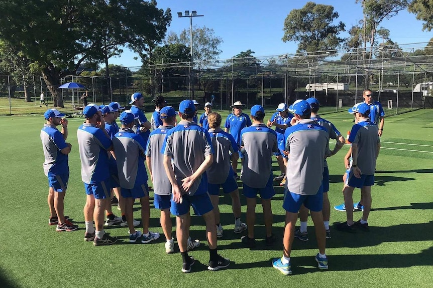 A group of men in blue and grey shirts stand in a group at the cricket nets.