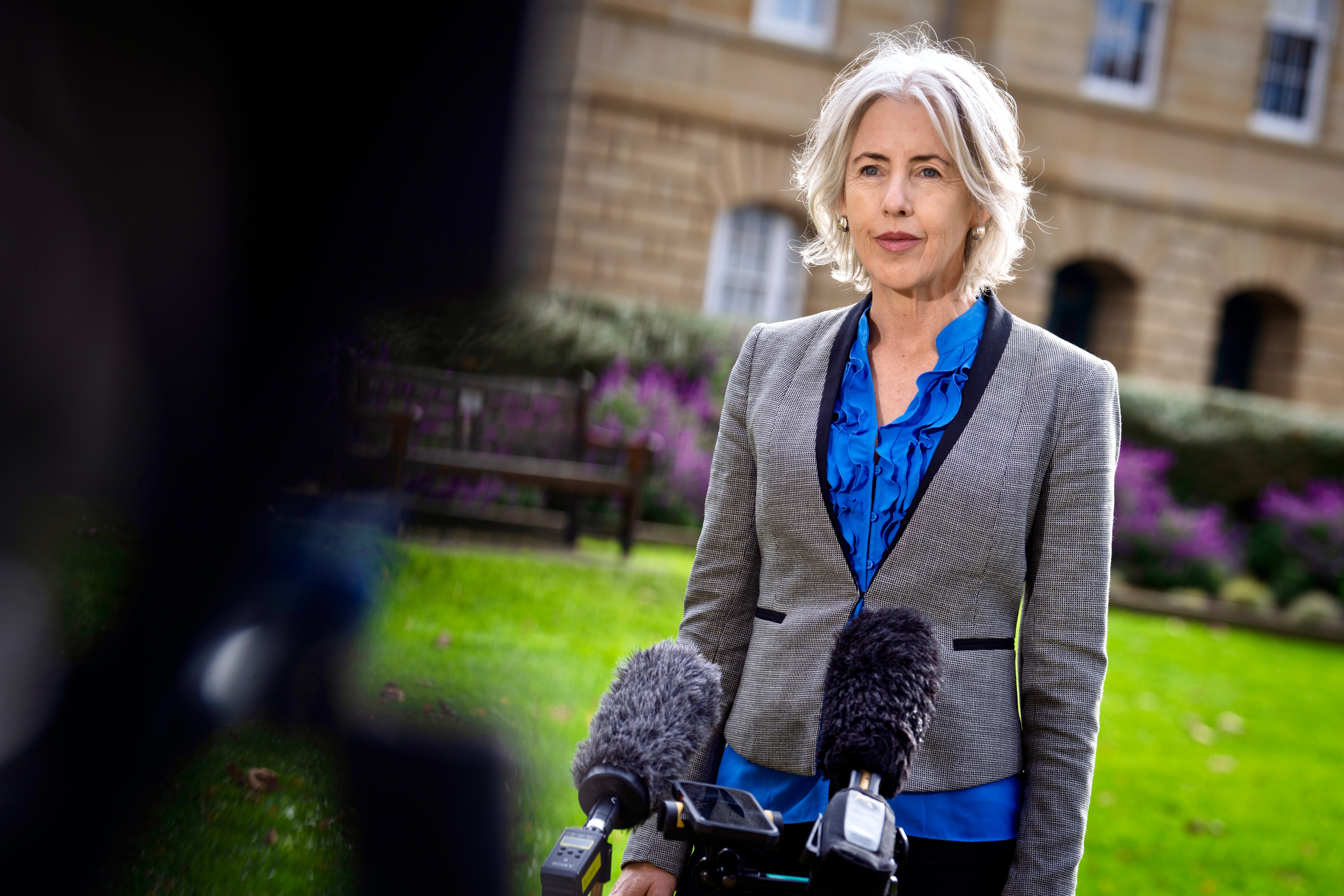 Rosalie Woodruff stands behind two television camera microphones with Parliament House in the background.