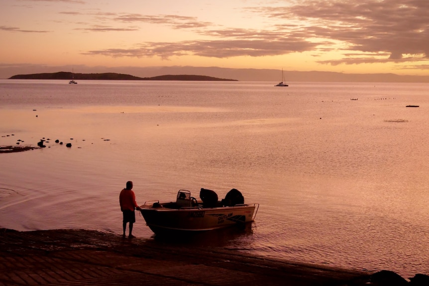 A man in silhouette stands near a boat on a beach, looking out to sea at during a pink and gold sunset.