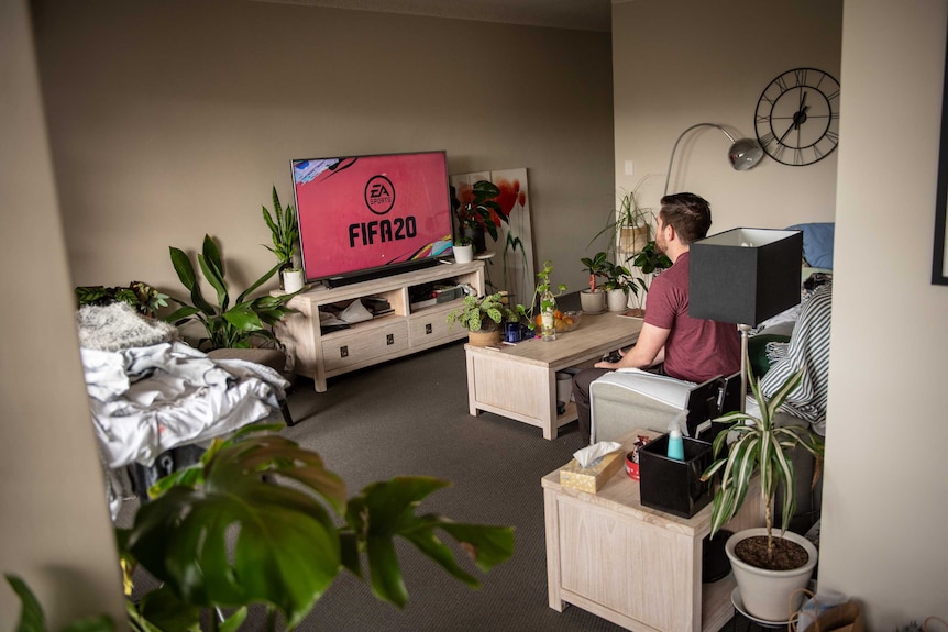 A young man sits on a couch in his apartment and plays FIFA20 on his PlayStation, surrounded by green plants.