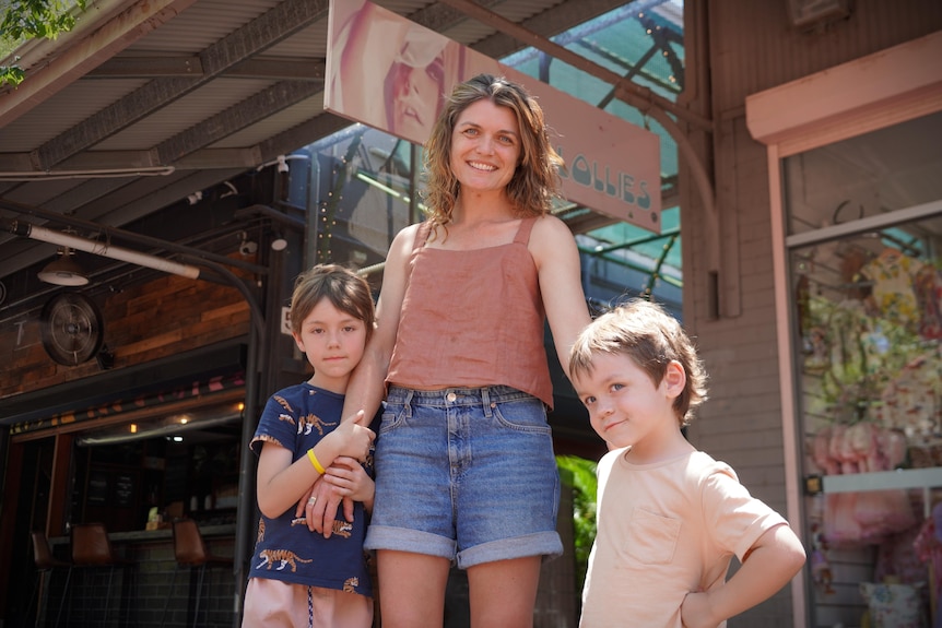 A woman with two young childen on either side of her, all smiling, in front of a strip of shops.