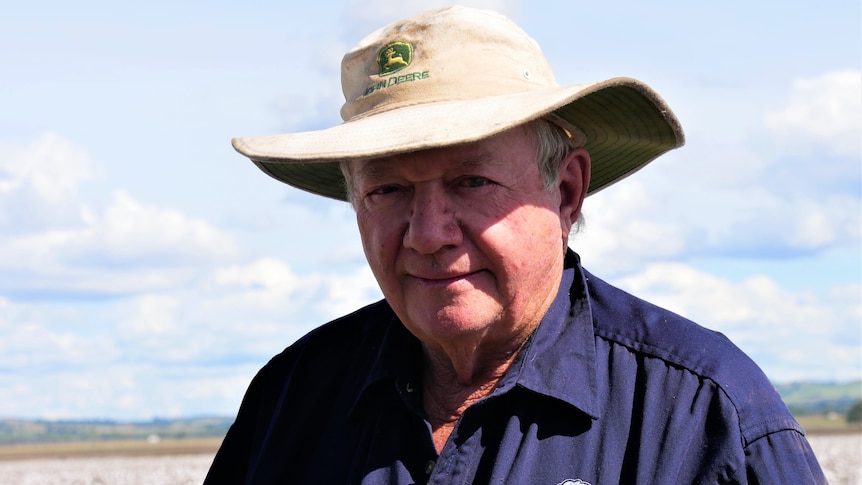 An older man in a dark blue work shirt and white broad brimmed hat standing in a cotton field. He seems hesitant.