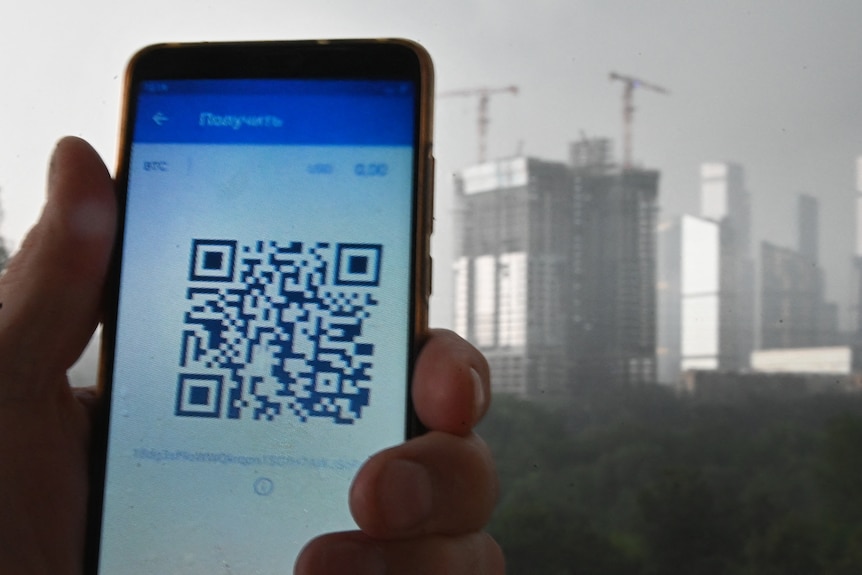 A left hand holds a mobile phone that is displaying a QR code, with tall buildings seen in the background.