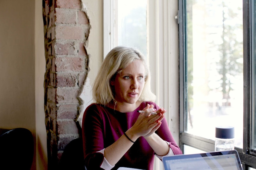 A woman sits looking out a cafe window with her hands together in front of her