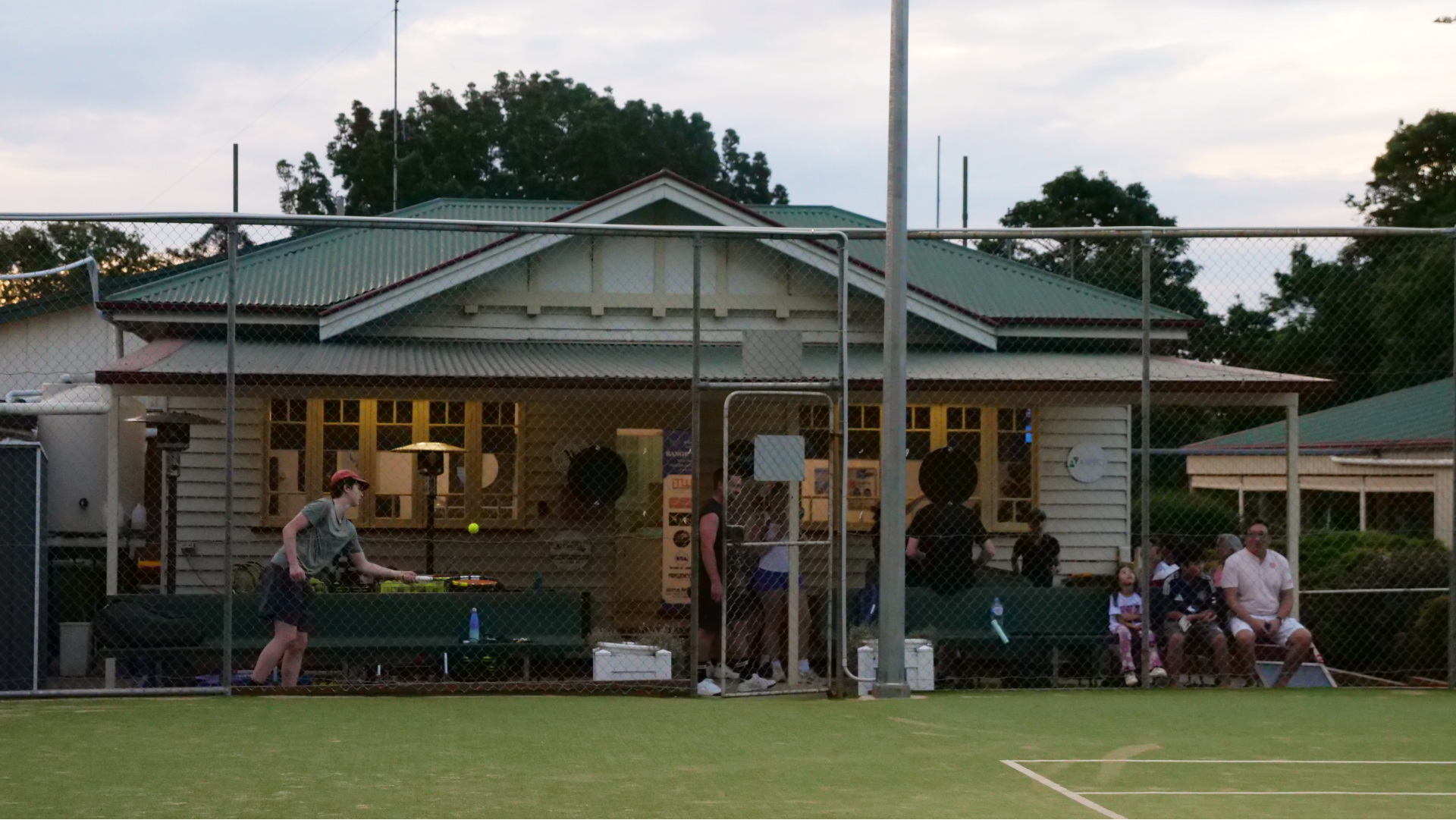 Una toma amplia de la casa club con gente sentada al frente y un adolescente haciendo rebotar una pelota en su bate.