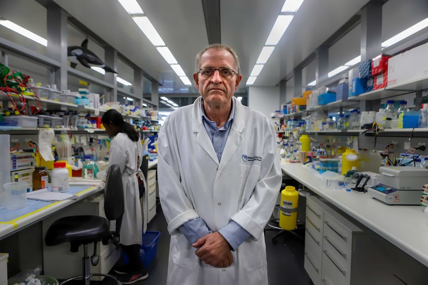 Man with glasses wearing lab coat stands in science laboratory with scientist behind him