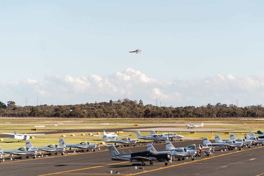 A wide shot showing a light plane in the sky above a runway with more than a dozen other aircraft parked on it.