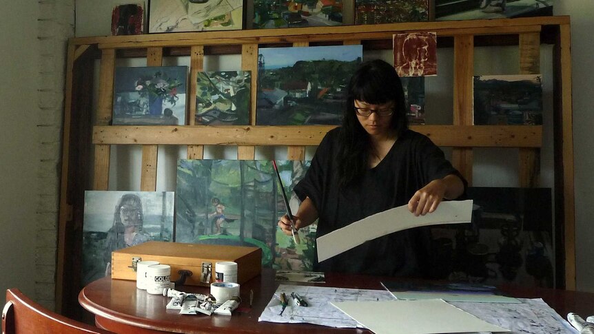 Anh Nguyen concentrating on her artwork in her studio, painting line the wall behind her