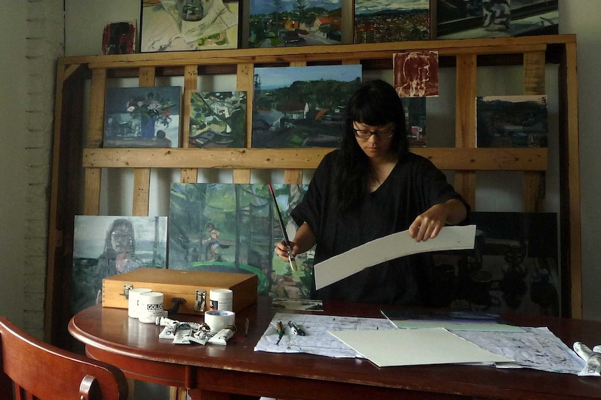 Anh Nguyen concentrating on her artwork in her studio, painting line the wall behind her