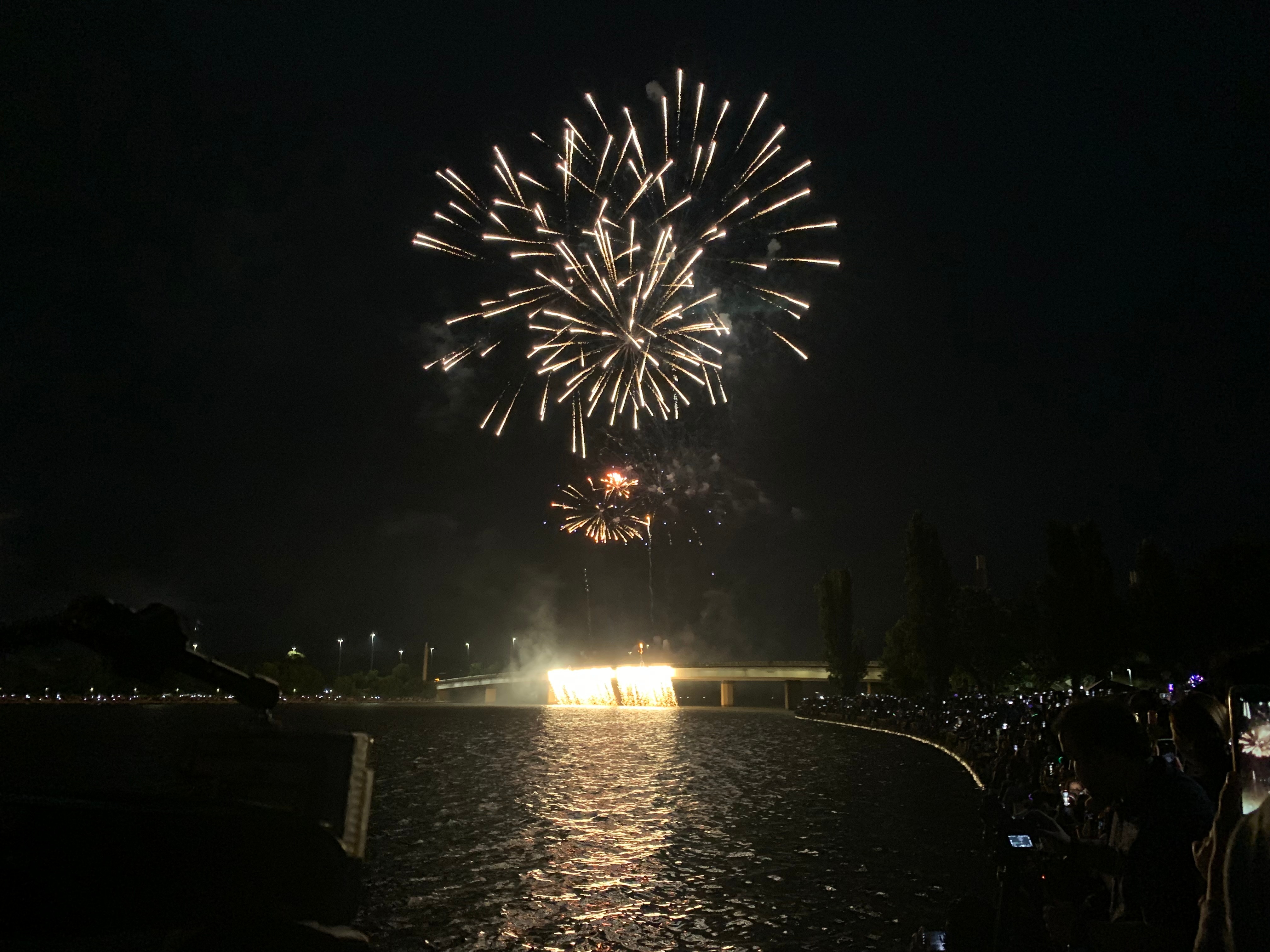 Una toma amplia de un espectáculo de fuegos artificiales nocturno sobre un puente en el lago Burley Griffin. Algunos fuegos artificiales fluyen desde el costado del puente.