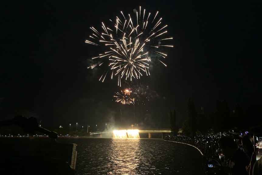 A wide shot at night of fireworks above a bridge on Lake Burley Griffin. Some fireworks flowing off the side of the bridge.
