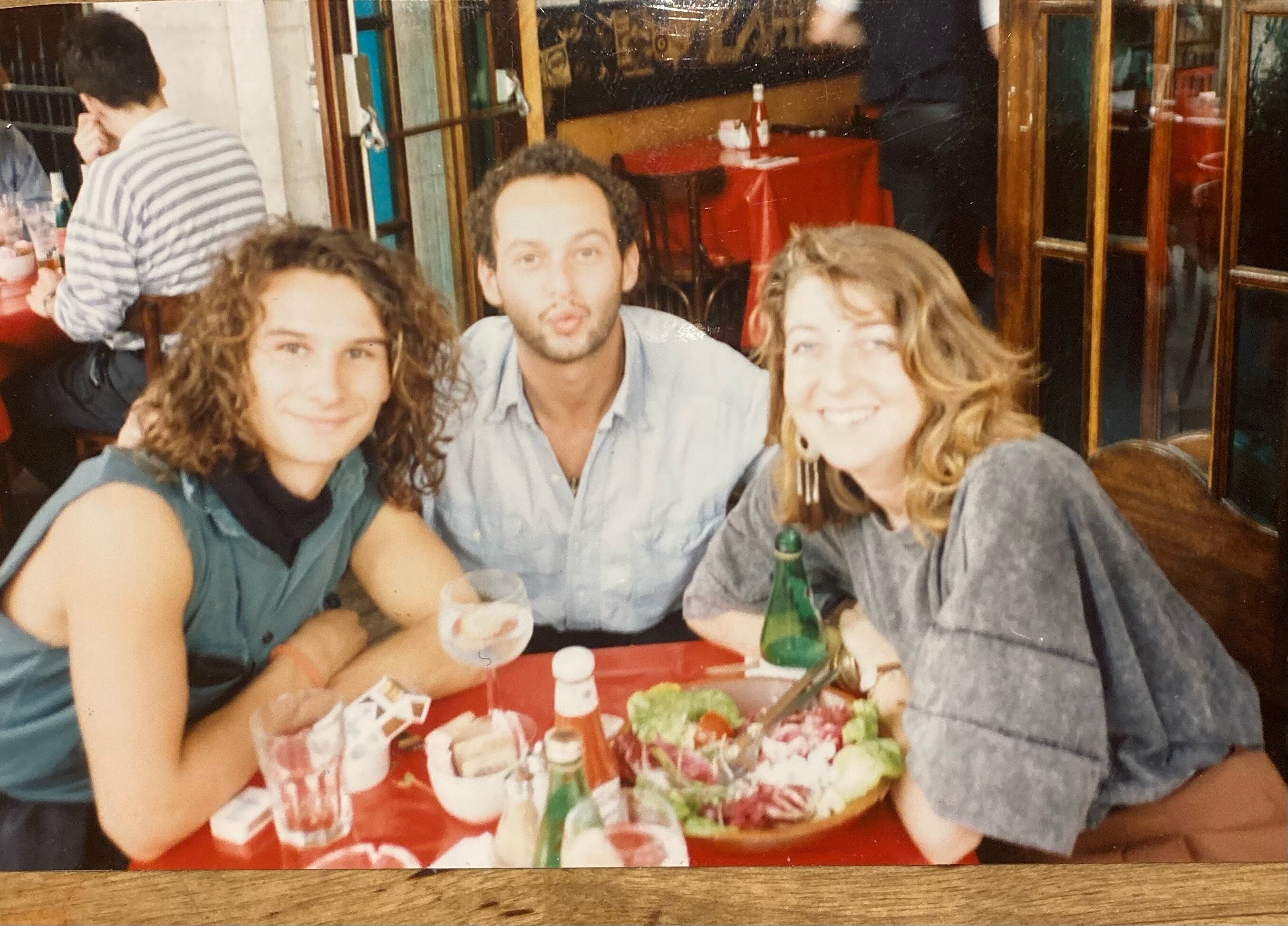 Three young people sit at a restaurant table smiling in a slightly blurry film photograph. 