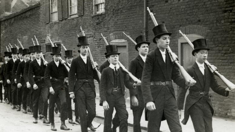 A black and white photo shows two straight rows of Eton students in top-hats carrying rifles marching down a street.