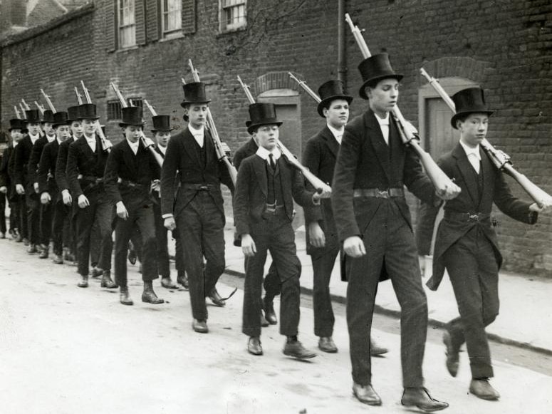 A black and white photo shows two straight rows of Eton students in top-hats carrying rifles marching down a street.