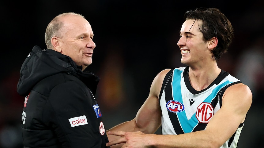 A smiling AFL coach holds hands with one of his players after a game.