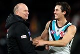 A smiling AFL coach holds hands with one of his players after a game.