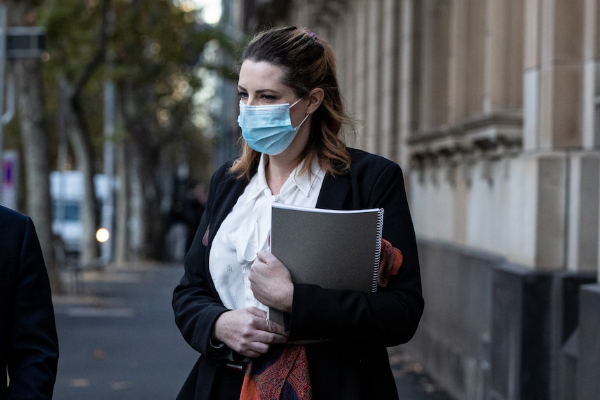 A woman holds a book of bound pages and wears a facemask as she walks down a city street