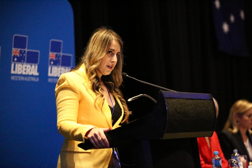 A woman wearing a yellow blazer speaks at a lectern.