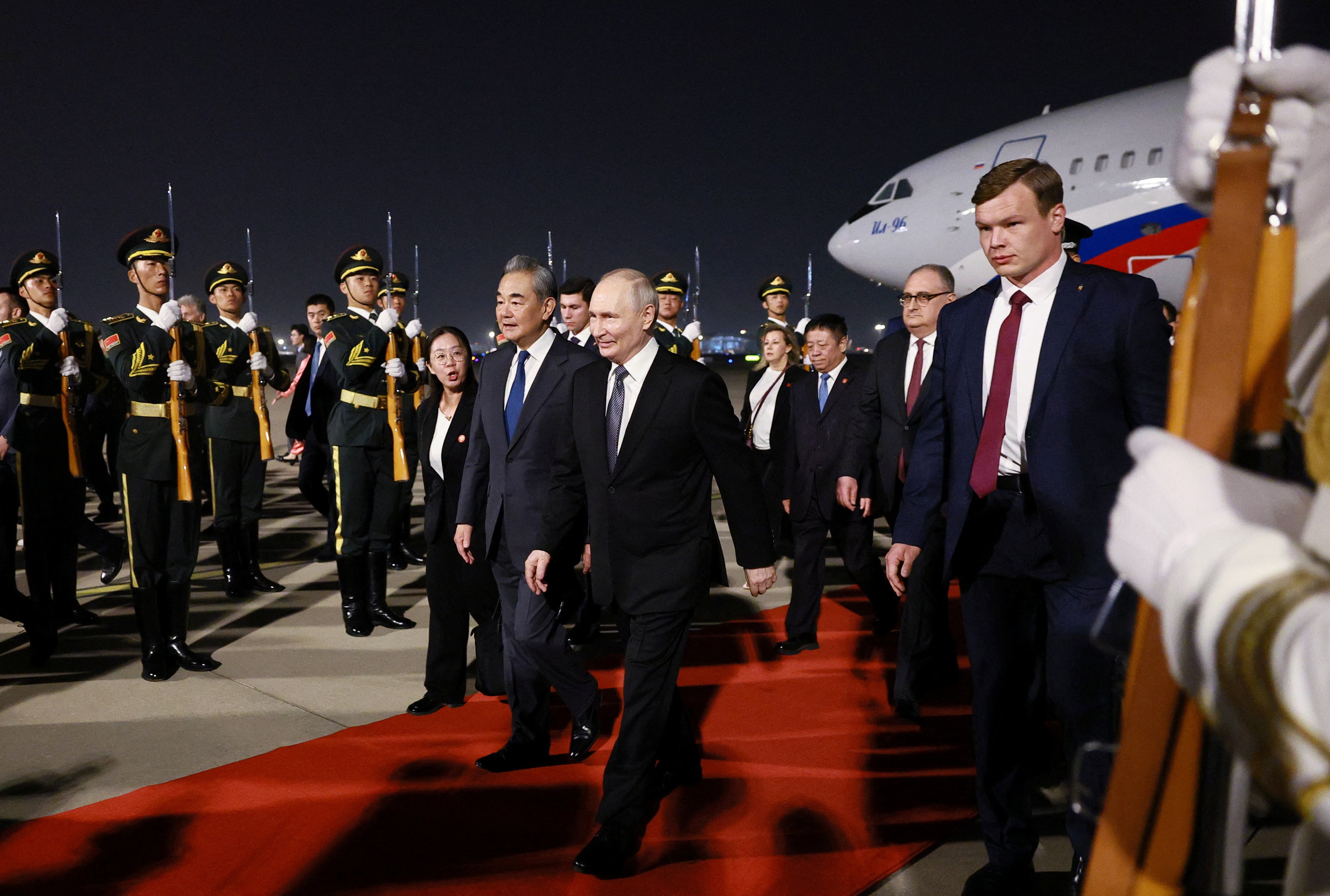 Two men walk side by side on a red carpet with the military creating a guard of honour.