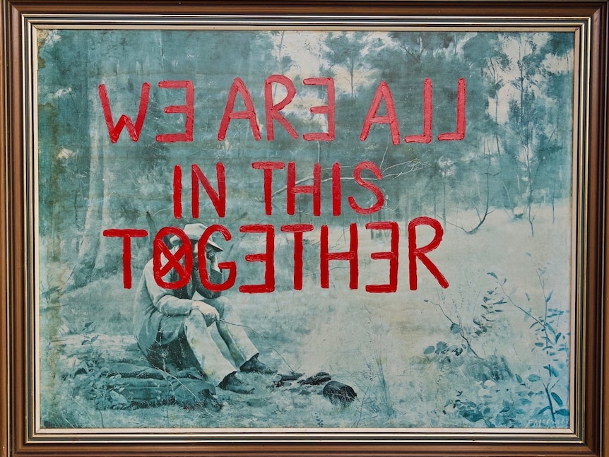 A framed print of a painting with the words 'We are all in this together' written in red across its surface.