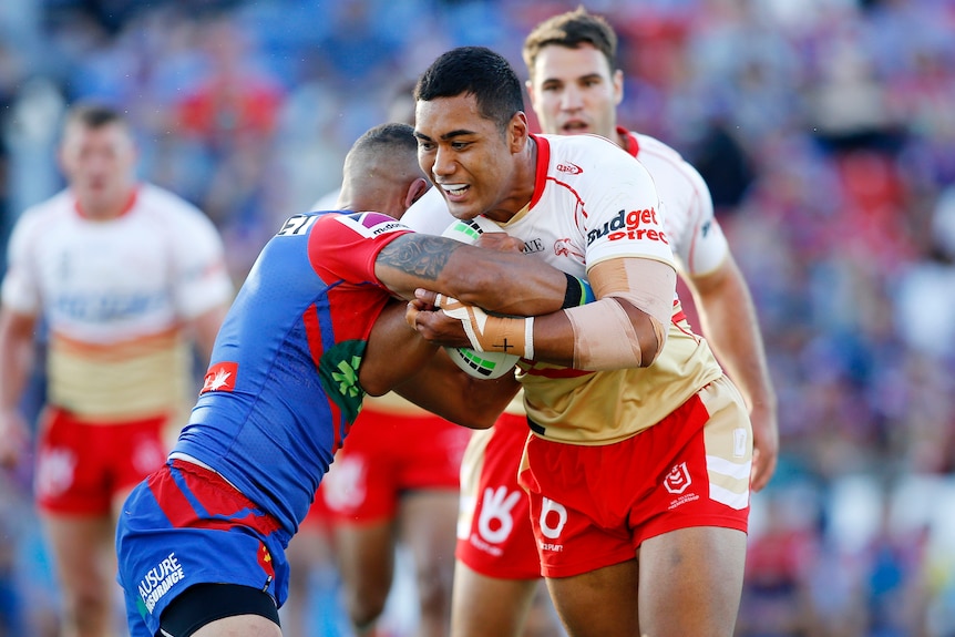 A Dolphins NRL player carries the ball while being tackled by a Newcastle Knights opponent.