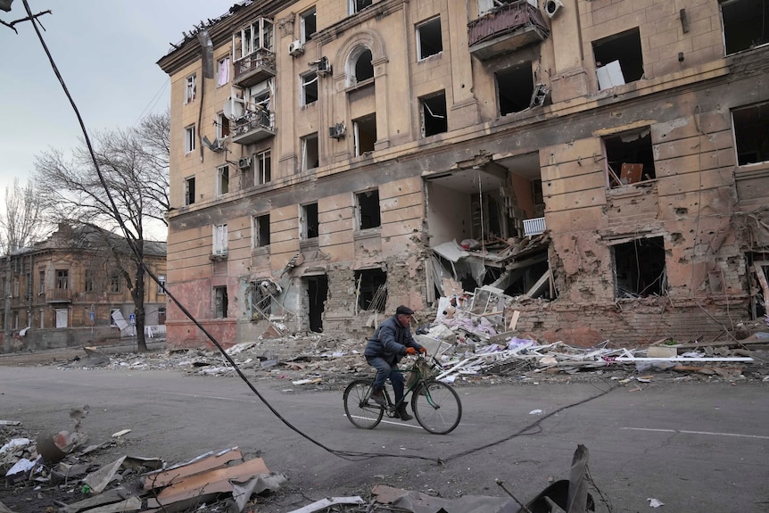 A man rides a bicycle on a road next to a large building which has been severely damaged.