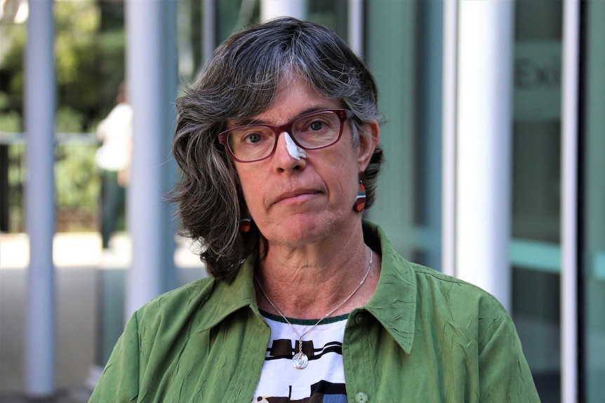 A woman with red glasses and a green shirt stairs directly into the camera and is not smiling