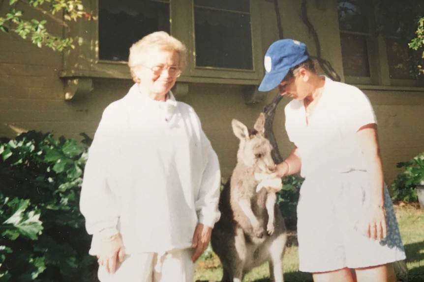 Two women wearing white with a small kangaroo between them.