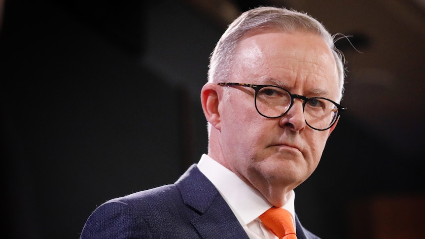 Albanese looks serious while wearing glasses and red tie, staring at the distance with a strong backlight framing him.