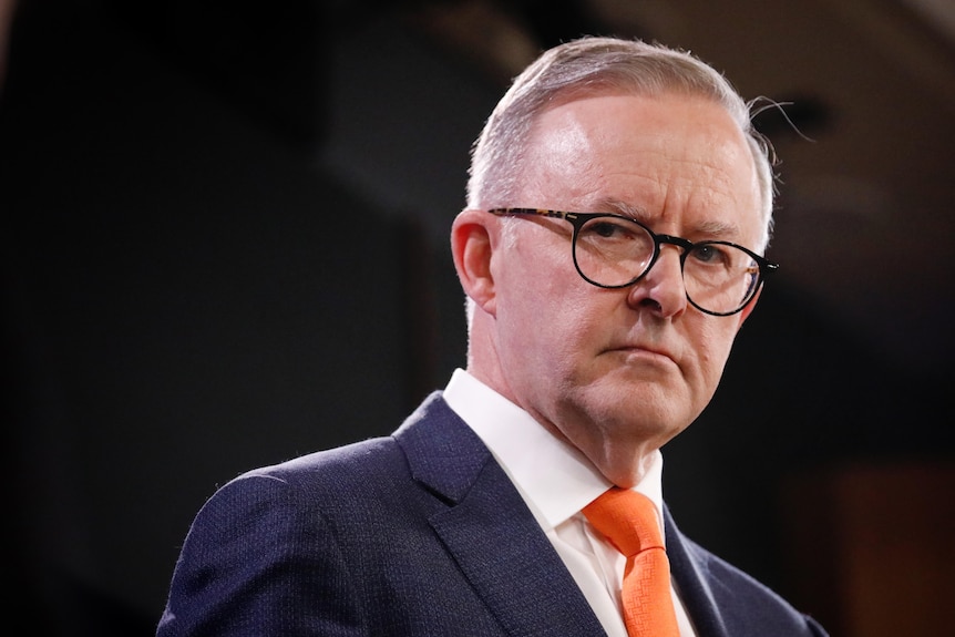 Albanese looks serious while wearing glasses and red tie, staring at the distance with a strong backlight framing him.