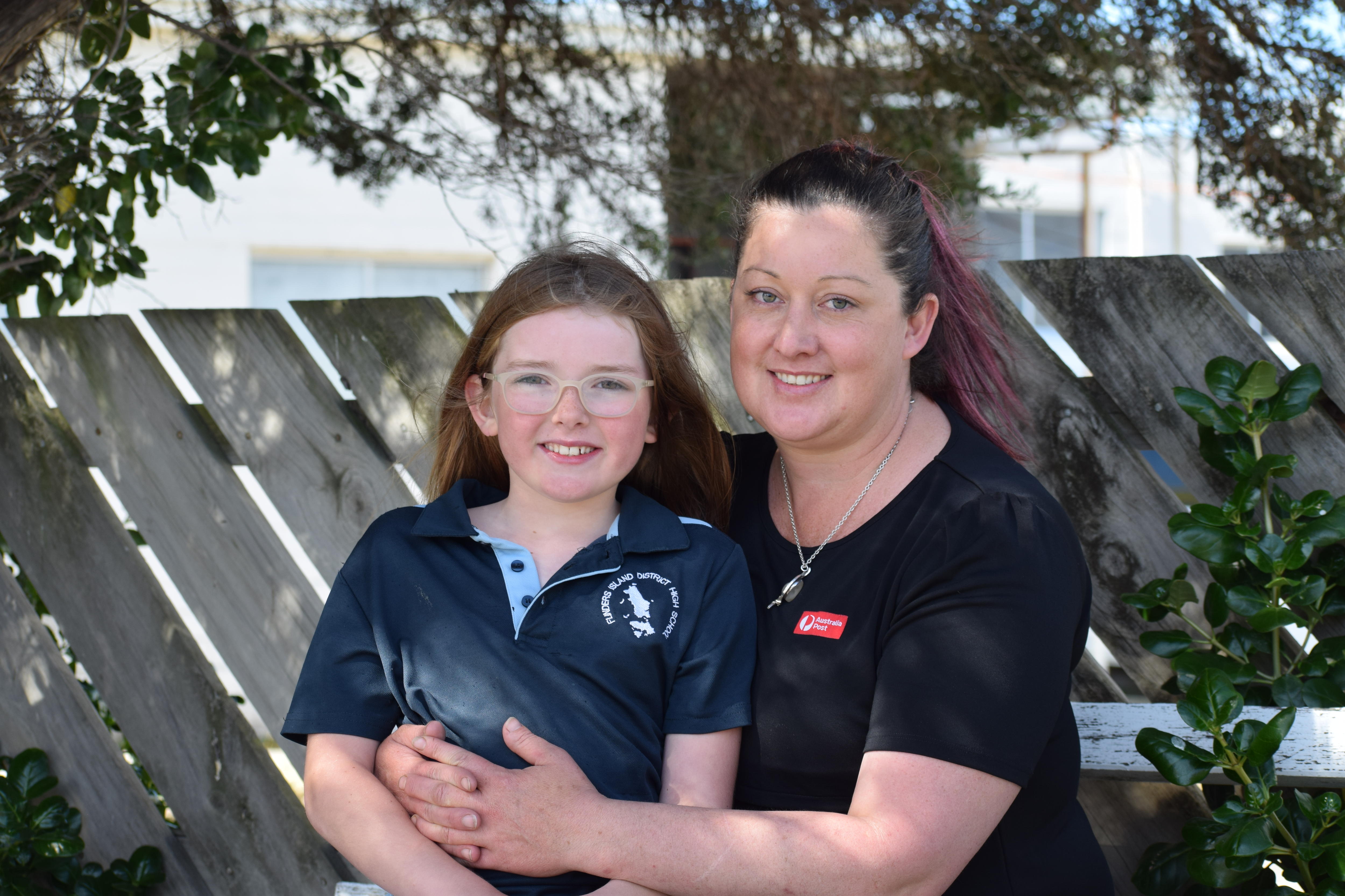 A woman sits with arms wrapped around girl. Girl wears glasses and is smiling.