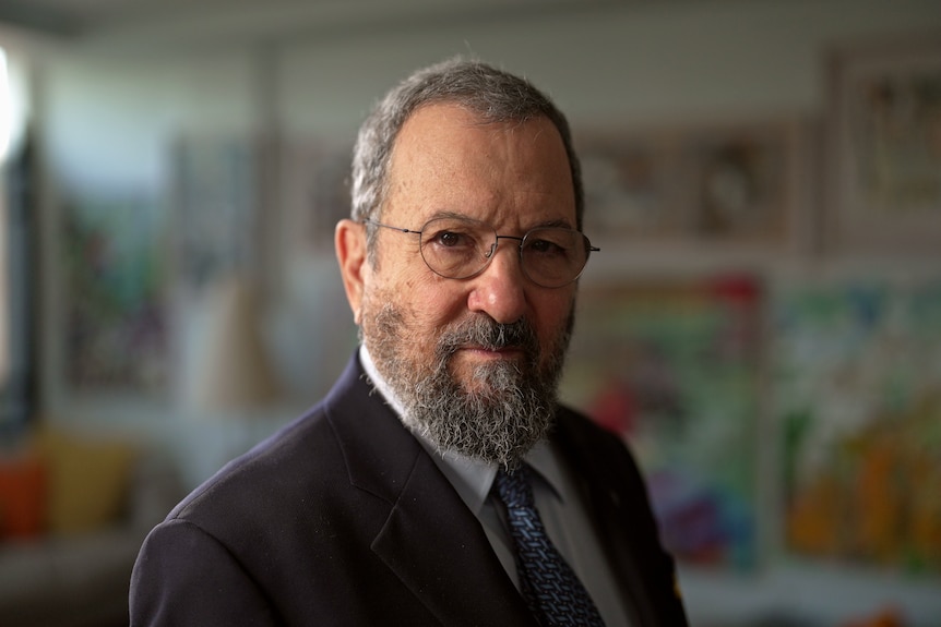 A man with glasses and a beard, wearing a suit, stands in a room, looking at the camera with a neutral expression.