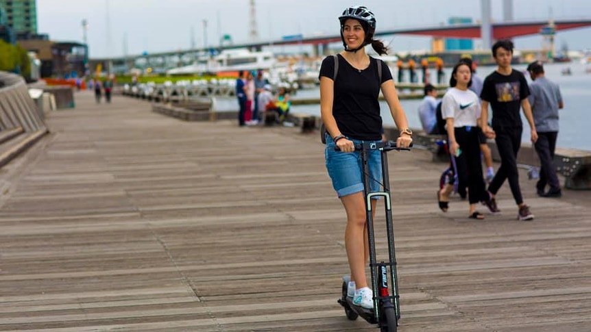 Michelle Mannering rides an e-scooter on a wharf wearing a helmet and smiling on a grey day.