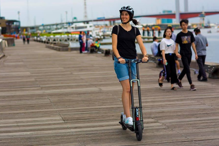 Michelle Mannering rides an e-scooter on a wharf wearing a helmet and smiling on a grey day.