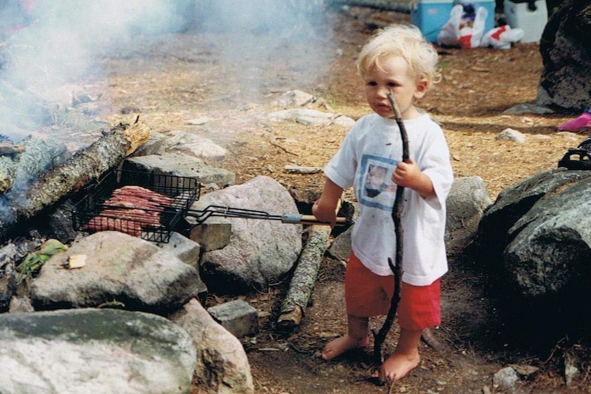 An old photo of a young blonde-haired boy holding a pan handle with meat over a fire.